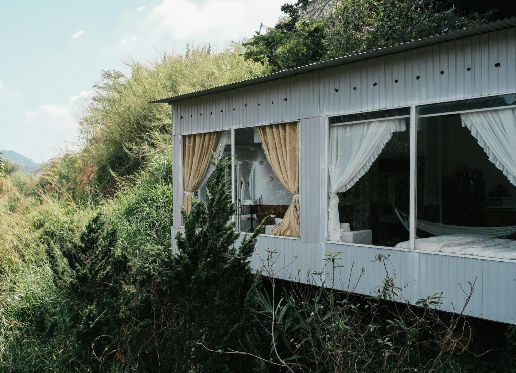 A serene glass house with open curtains amidst dense foliage under a bright daytime sky.