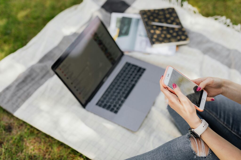 From above crop anonymous female student in blue jeans text messaging on smartphone while sitting on blanket with laptop and college books in green park