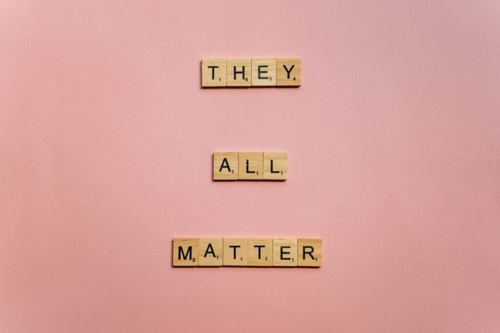 Wooden Scrabble tiles spell 'They All Matter' on a pink background, promoting inclusivity.